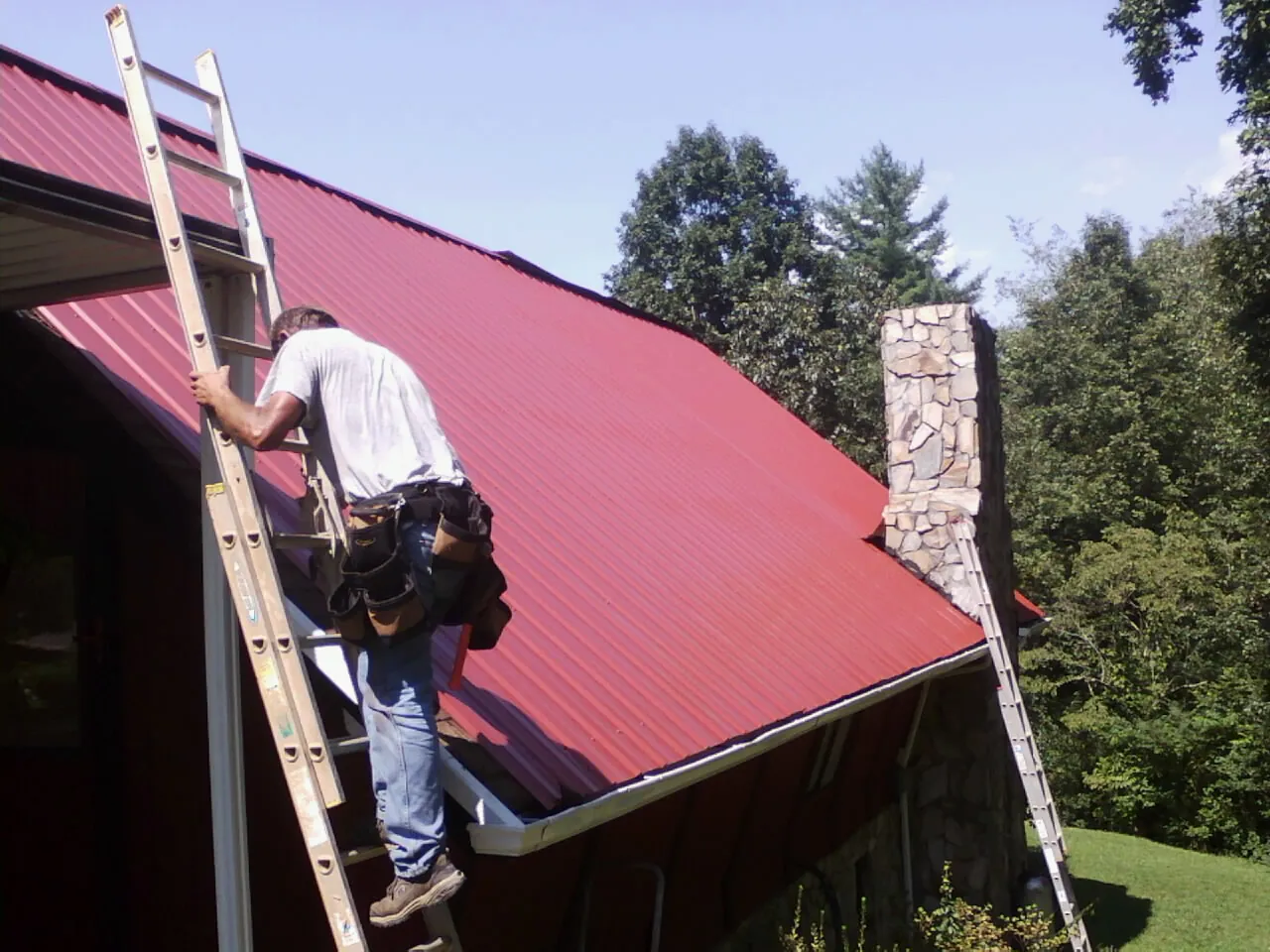 Roofer on a ladder beside a red metal roof and stone chimney — Soffit & Fascia Repair by Roofing Brothers in Southbury